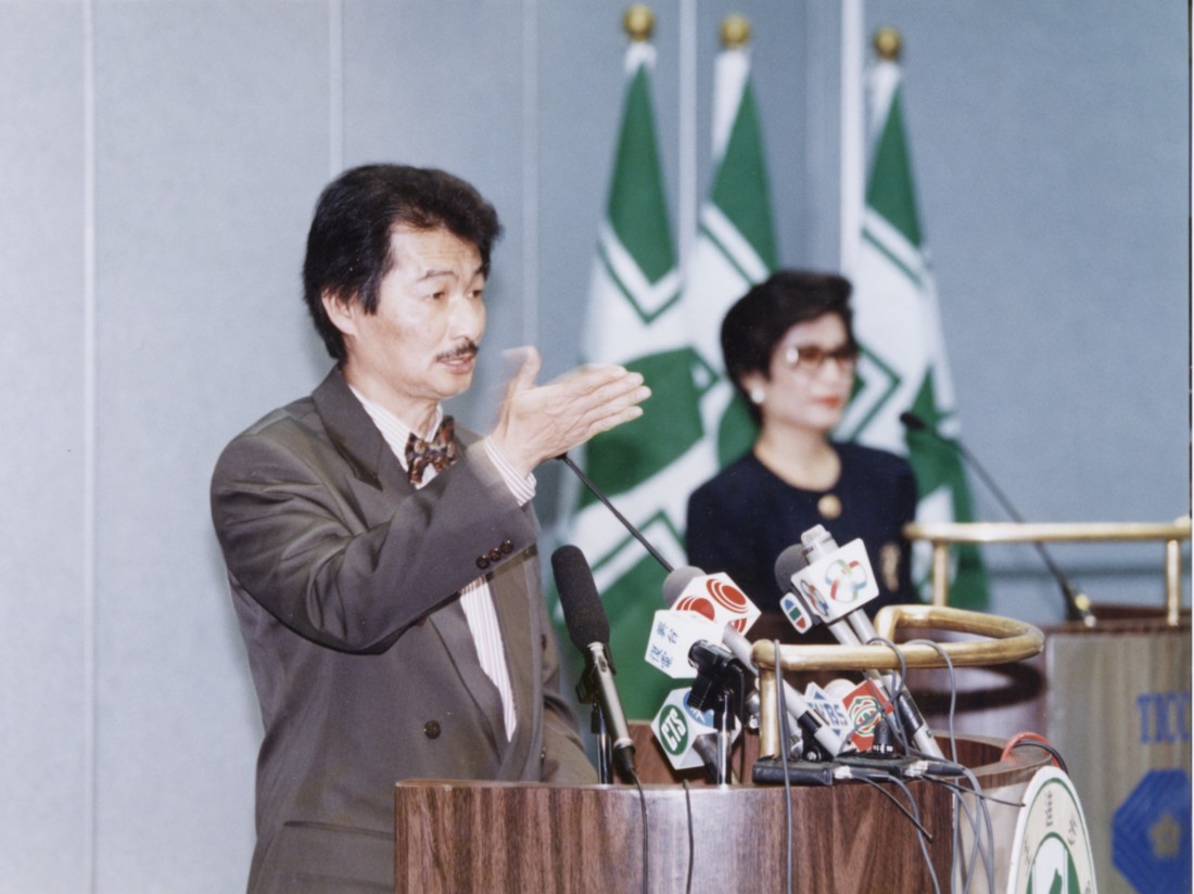 Man in a suit speaks at a wooden podium during a press conference, gesturing with his hand while microphones sit before him; green-and-white flags in the background.