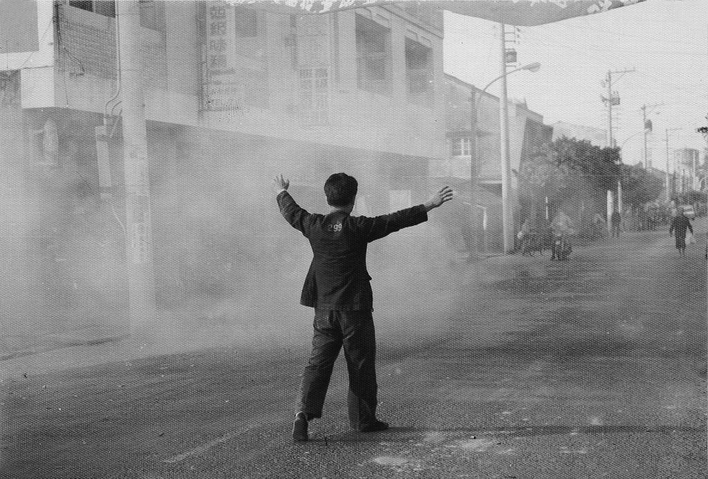 Man standing in the middle of a smoky street with arms raised, facing away from the camera, buildings lining the road.