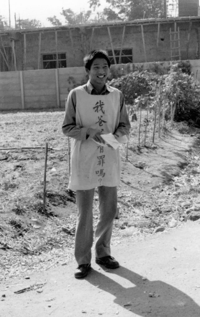 Man standing outdoors, wearing a cloth sign with Chinese text, smiling at the camera; text appears to read '我會有罪嗎?' (Am I guilty?).