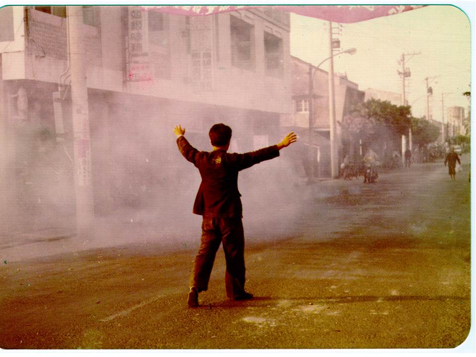 Man in a dark suit stands with arms raised in a dusty, smoky city street, storefronts on both sides behind him.