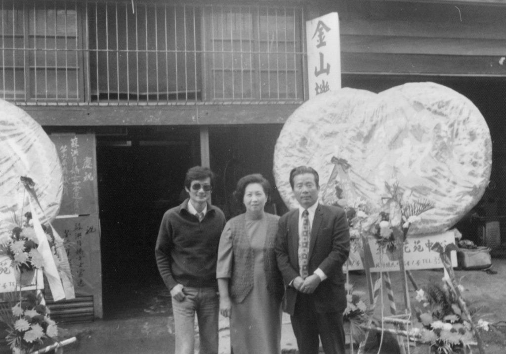 Three people stand together outside a building with large round floral wreaths, likely at a grand opening or celebration event (black-and-white photo).
