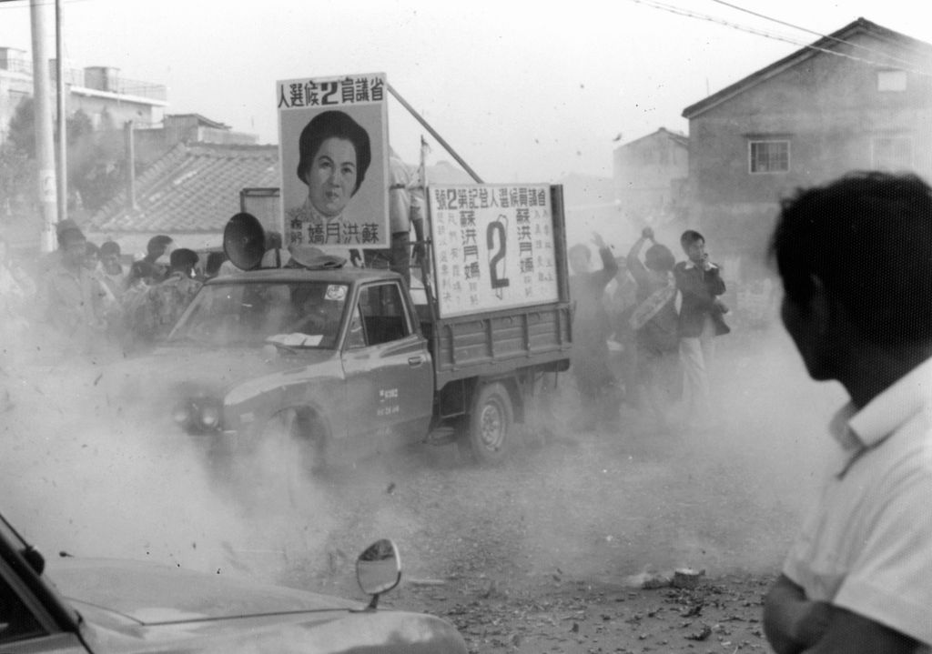 Campaign truck with large portrait posters leading a dusty street crowd in a political rally (Chinese signage visible).