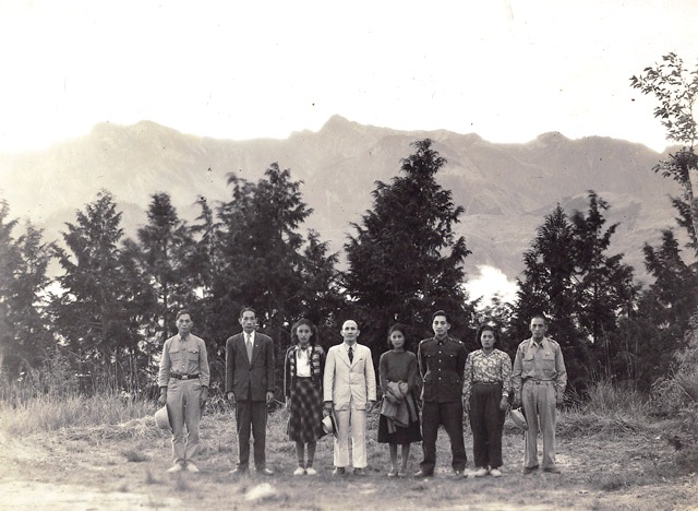 Group of nine people standing in a line outdoors, with trees and mountains in the background (mixed formal and military attire).