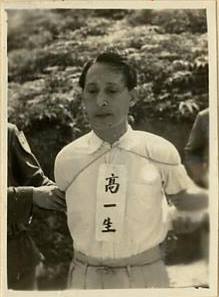 Black-and-white photo of a man in a white shirt with a vertical strip of characters pinned to his chest, standing outdoors with others slightly visible in the background.