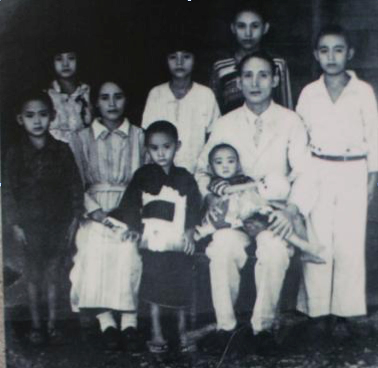 Black-and-white family portrait with two adults seated center, a baby on the lap, and several children standing around them.