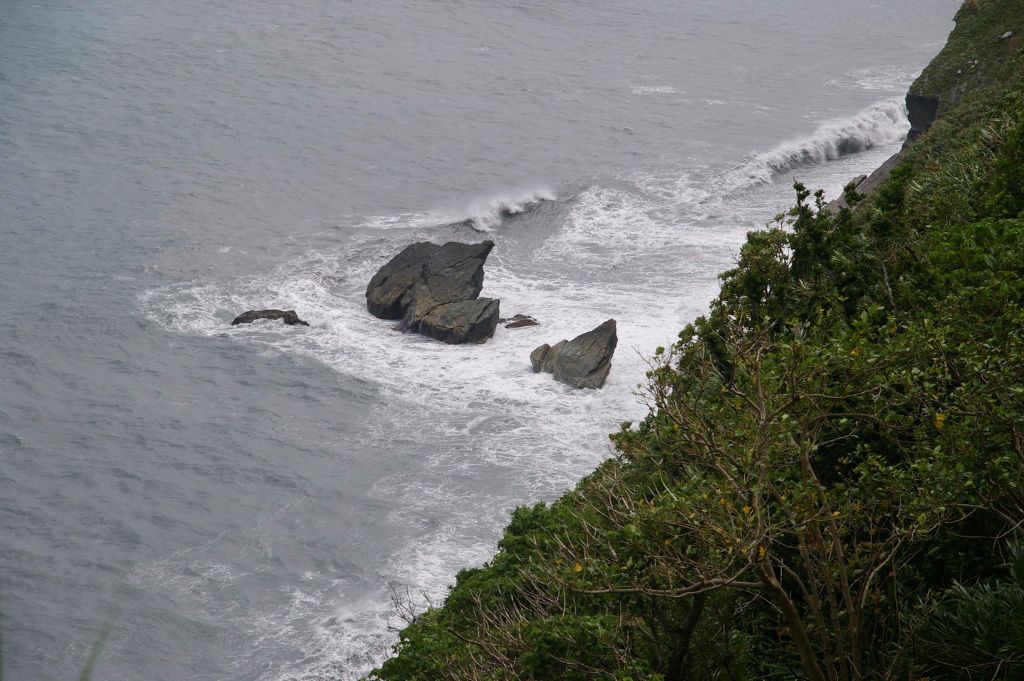 Cliffside view of the rocky shoreline with waves crashing against large dark rocks, and green vegetation on the right.
