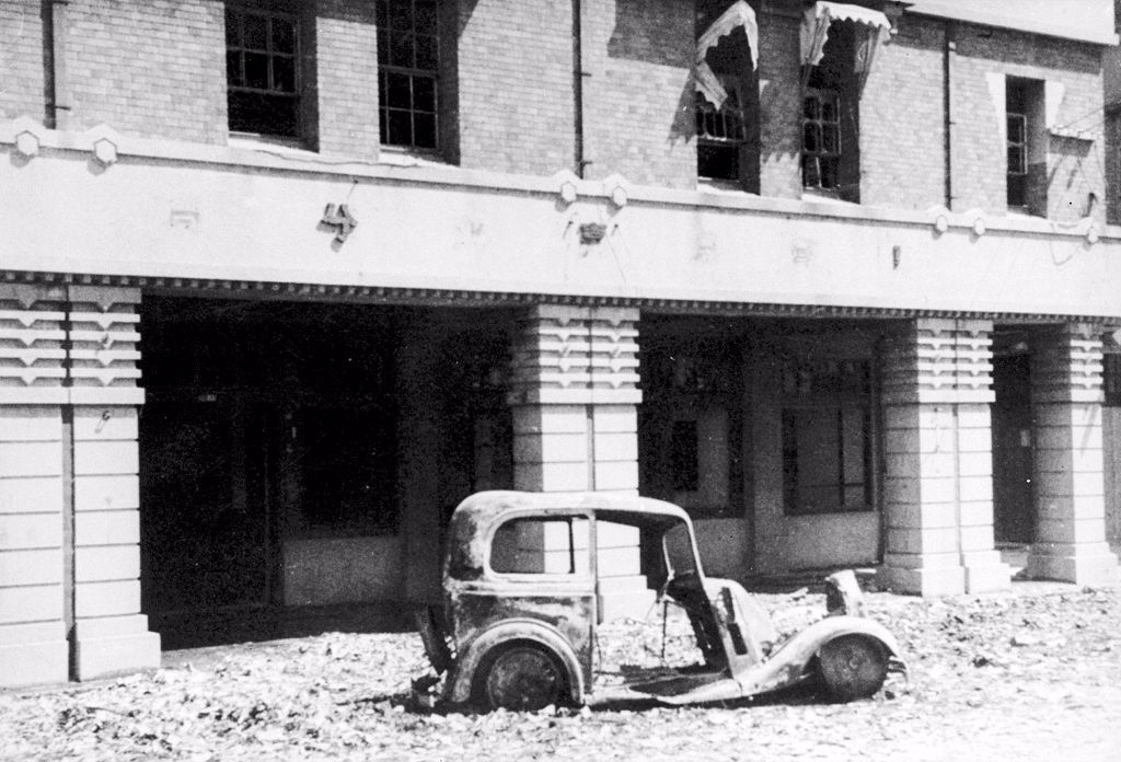 Abandoned, rusted vintage car shell on rubble-strewn street in front of a war-damaged building with columns and arched storefronts.