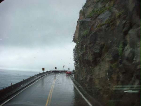 Windshield view of a rainy coastal highway: a slick, reflective road curves along a cliff to the right with the sea on the left; a small red car is ahead in the distance.
