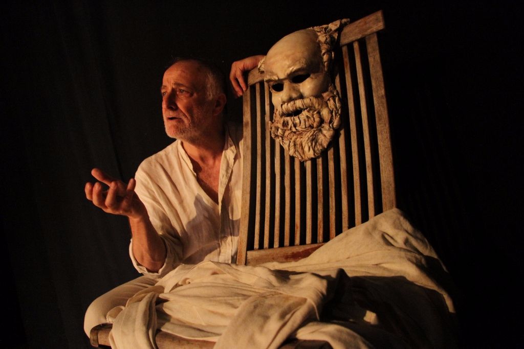 Older man in a white shirt sits beside a wooden chair draped with white fabric, a carved mask with a beard hangs on the chair, on a dark stage backdrop.