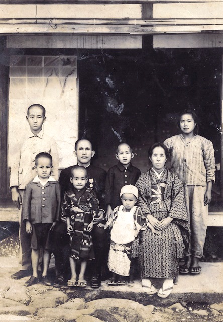 A multi-generational family portrait on a porch, with parents and several children in early 20th‑century clothing posed in front of a house.