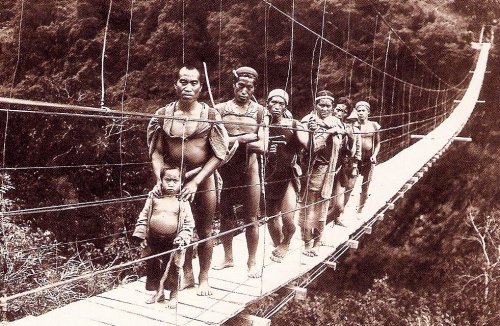 Indigenous people walking a narrow suspension bridge with a small child at the front over a forested gorge.