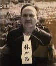 Black-and-white photo of a man standing outdoors, a paper tag with Japanese writing hangs from his neck.