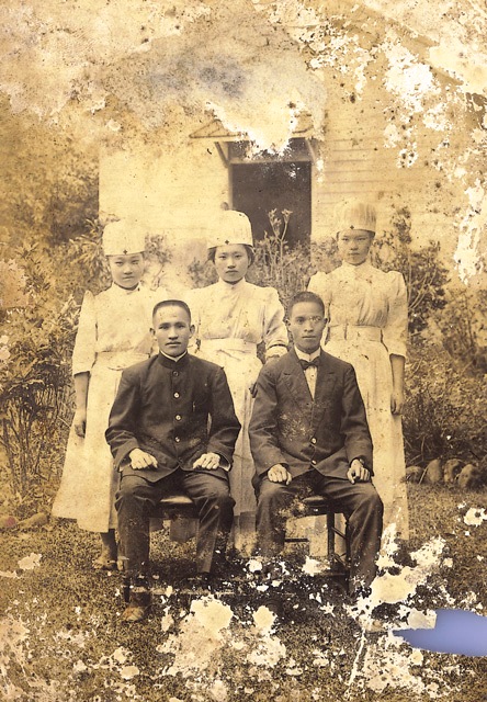 Sepia-era portrait of two men in suits seated in front, with three women in white dresses and nurse caps standing behind them outdoors.