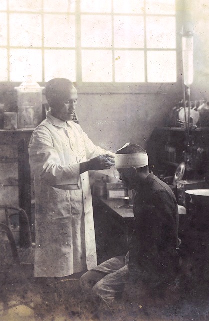 Old black-and-white photo of a doctor in a white coat applying treatment to a seated patient with a head bandage in a vintage clinic.