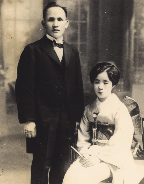 Historic studio portrait of a man in a suit standing beside a seated woman in a kimono.
