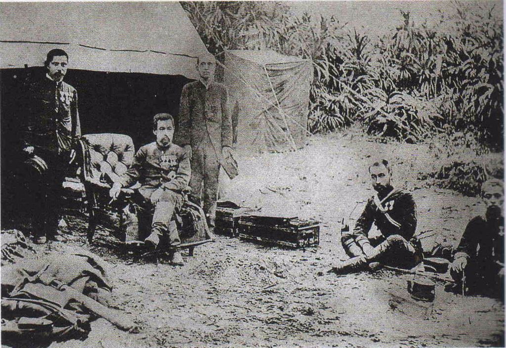 Soldiers in uniform sit around a canvas tent at a dusty camp, with crates and gear scattered nearby.