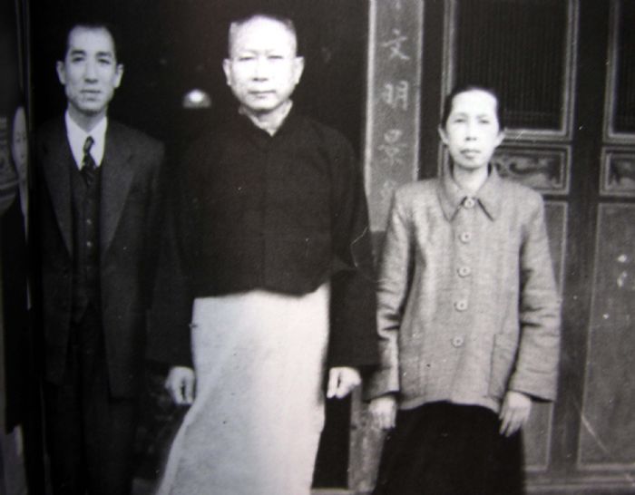 Three adults stand side by side in front of a doorway with Chinese characters, wearing formal clothing (left: man in suit, center: older man in traditional attire, right: woman in blazer).
