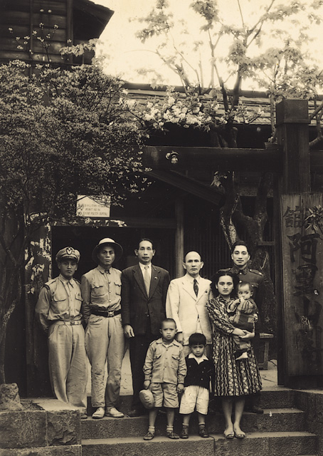 Sepia-toned group portrait: two soldiers in uniform, two men in suits, two women with children, standing on steps before a traditional wooden gate and building.