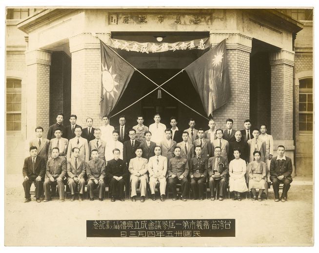 Formal group portrait of about 40 East Asian men and a few women in suits and uniforms, posed on the steps outside a brick building with two crossed flags behind them and a plaque in front.