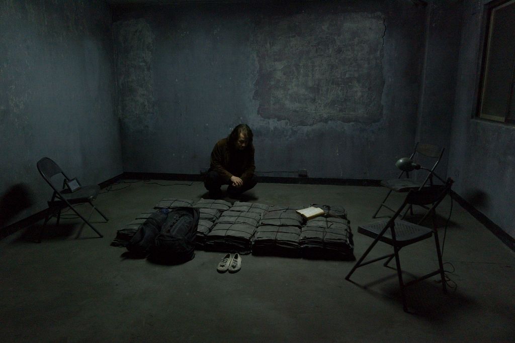 A person sits cross‑legged on the floor in a dim, gray basement-like room, facing a grid of stacked black bags with a backpack, a notebook, and shoes nearby. The scene feels somber and isolation‑like.