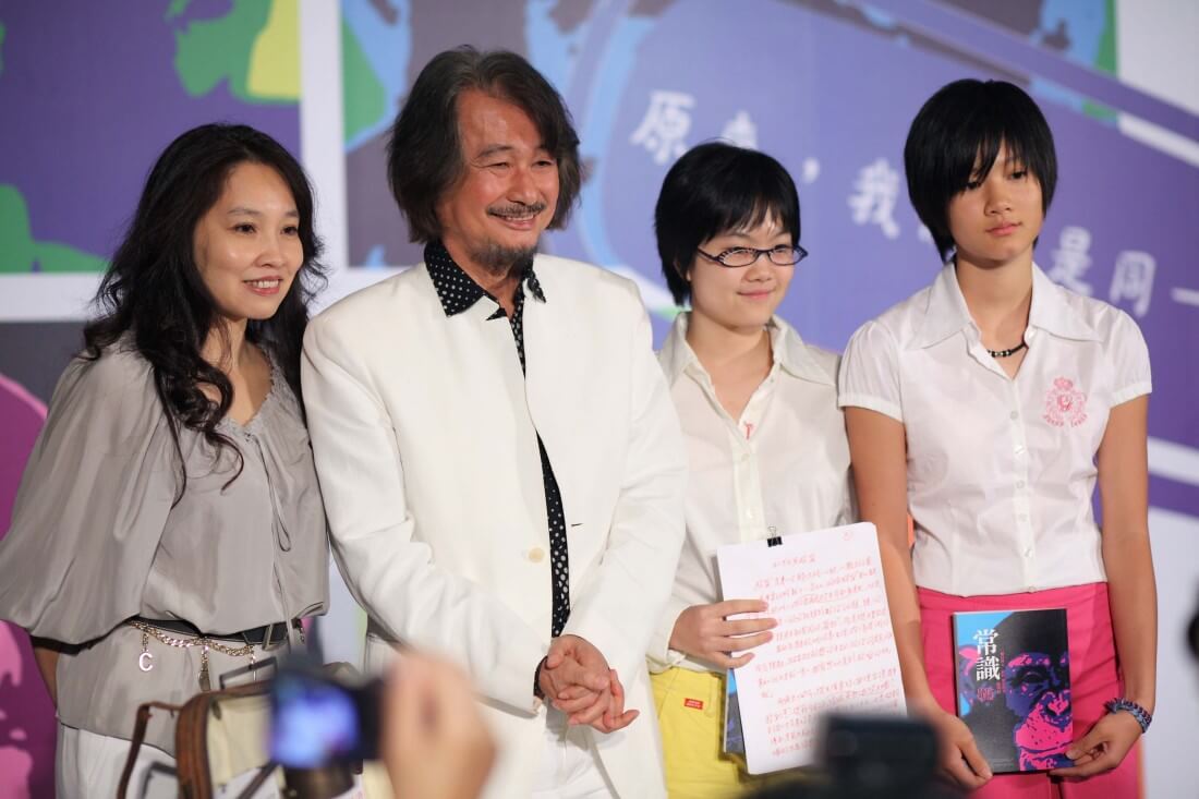 Four people posing on a stage: a woman in gray on the left, a man in a white suit center, and two girls in white shirts holding papers beside him.
