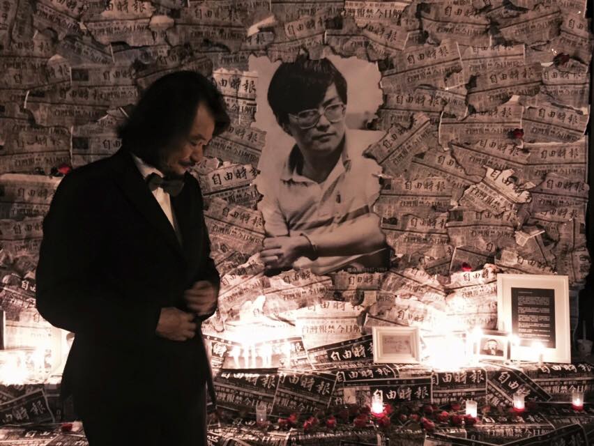Man in a suit adjusting his jacket in a memorial installation with a wall covered in newspapers and a large portrait of a woman, candles and plaques nearby