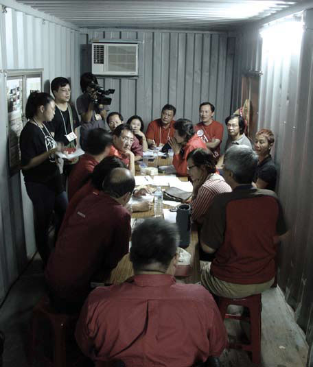 Group of people gathered around a long table in a small metal-walled room, some wearing red shirts, with a camera crew filming nearby.