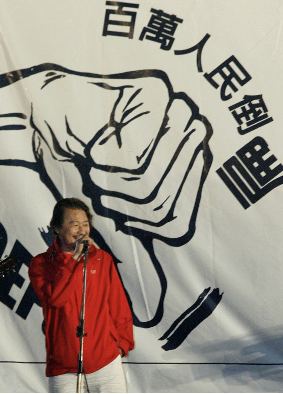 Woman in a red jacket singing into a microphone on a stage with a large black-and-white fist illustration as backdrop.