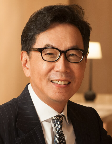 Asian man in a dark pinstripe suit and patterned tie, wearing black-framed glasses, smiling at the camera indoors.