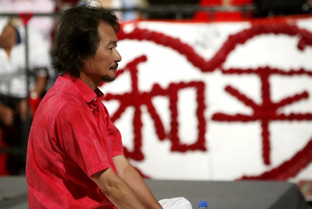 Profile of a man in a red shirt seated, facing left, with a red decorative banner behind him at an event.