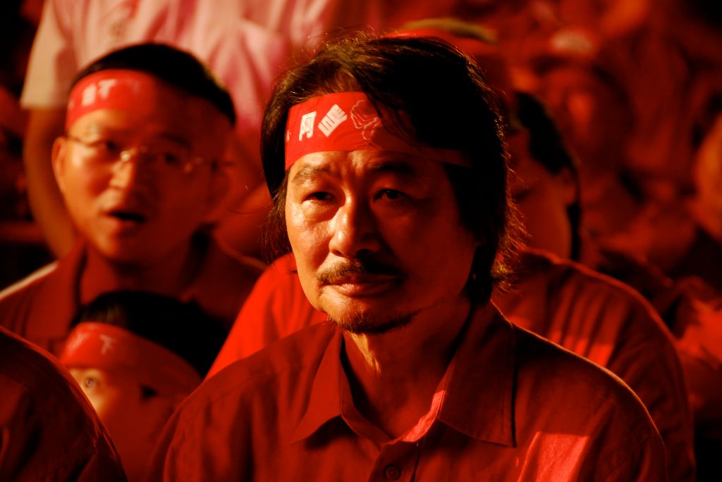Man with a red headband gazes forward in a crowded, red-lit gathering or protest.