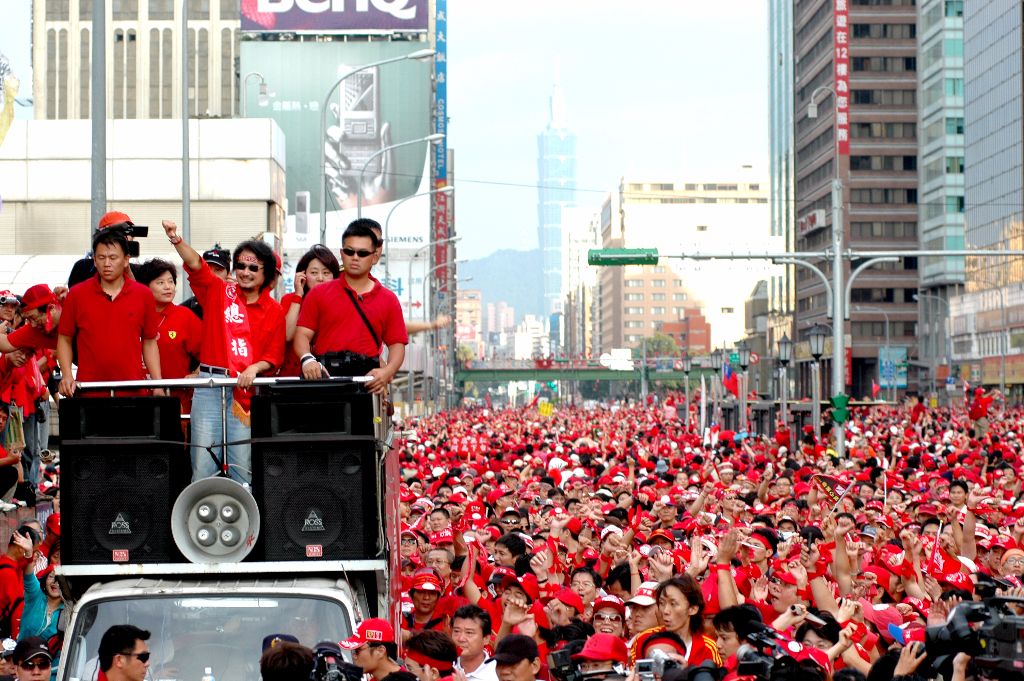 Thousands of people dressed in red fill a city street for a parade, fronted by a speaker truck; Taipei 101 is visible in the distant skyline.