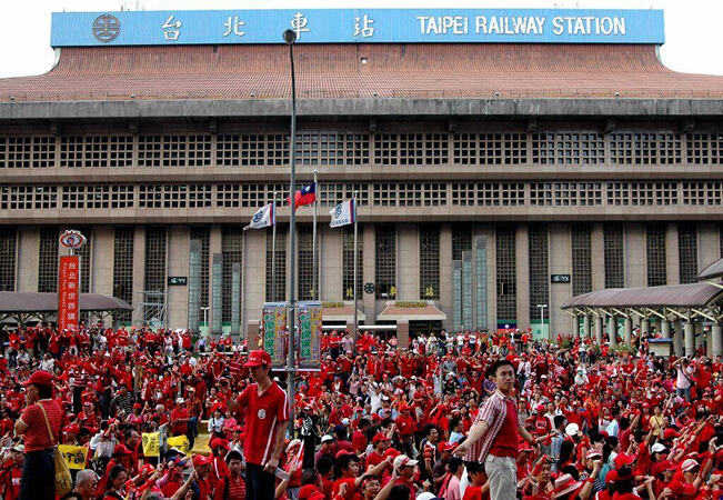 Crowd of people wearing red clothing gathered in front of Taipei Railway Station, signage visible at the entrance.