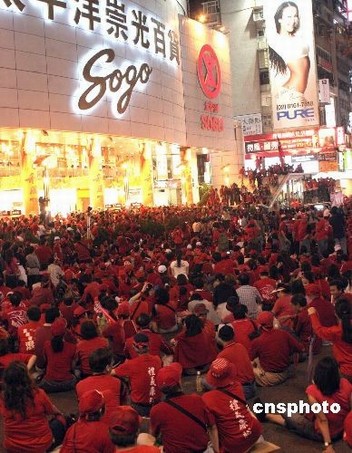 Large crowd of people wearing red shirts gathered outside a brightly lit department store at night, storefront signs and advertisements visible.
