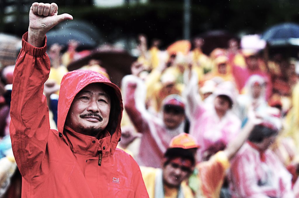 Man in a red rain jacket with hood raising his fist at a cheering crowd in the rain, umbrellas visible in the background.