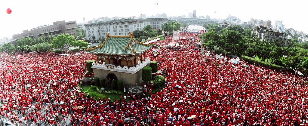 Aerial view of a massive crowd wearing red around a historic Chinese pavilion in a city square park area.