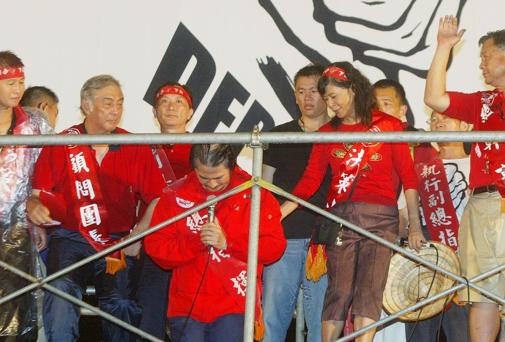 Group of people in red outfits and sashes on a stage with a metal railing, one man in front bowing and speaking into a microphone in his hand, others watch behind him.
