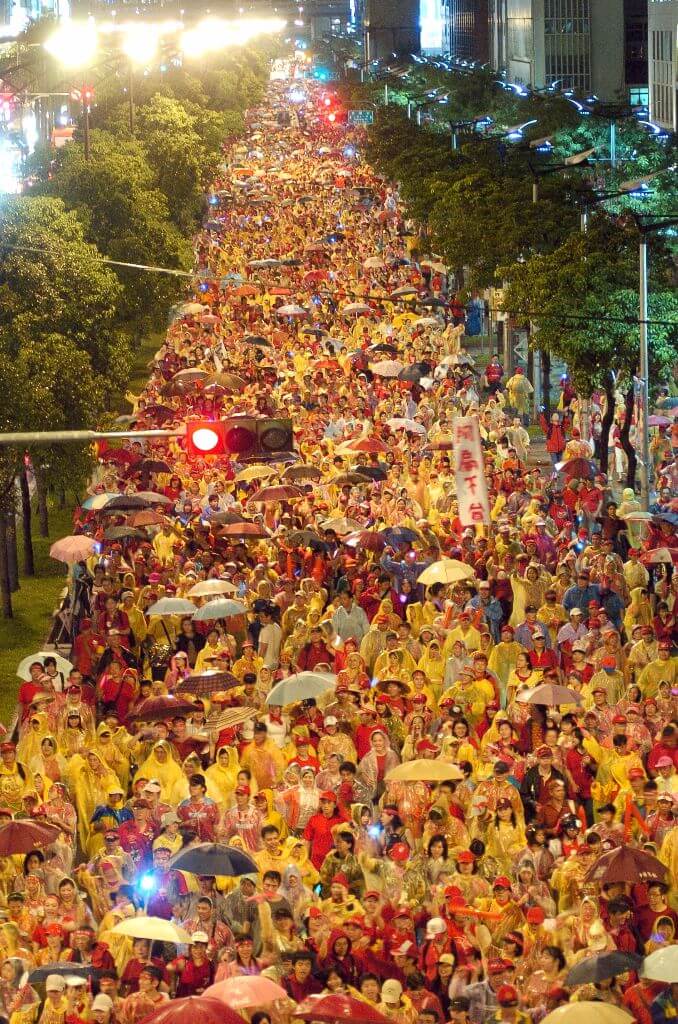 Massive crowd walking down a city street at night, many carrying umbrellas or wearing rain ponchos under bright lights.