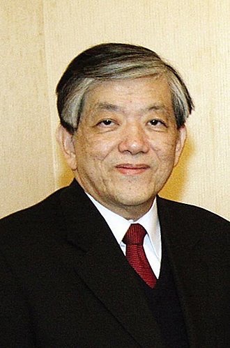 Portrait of an older Asian man in a dark suit, white shirt, and red tie, facing the camera against a beige background.