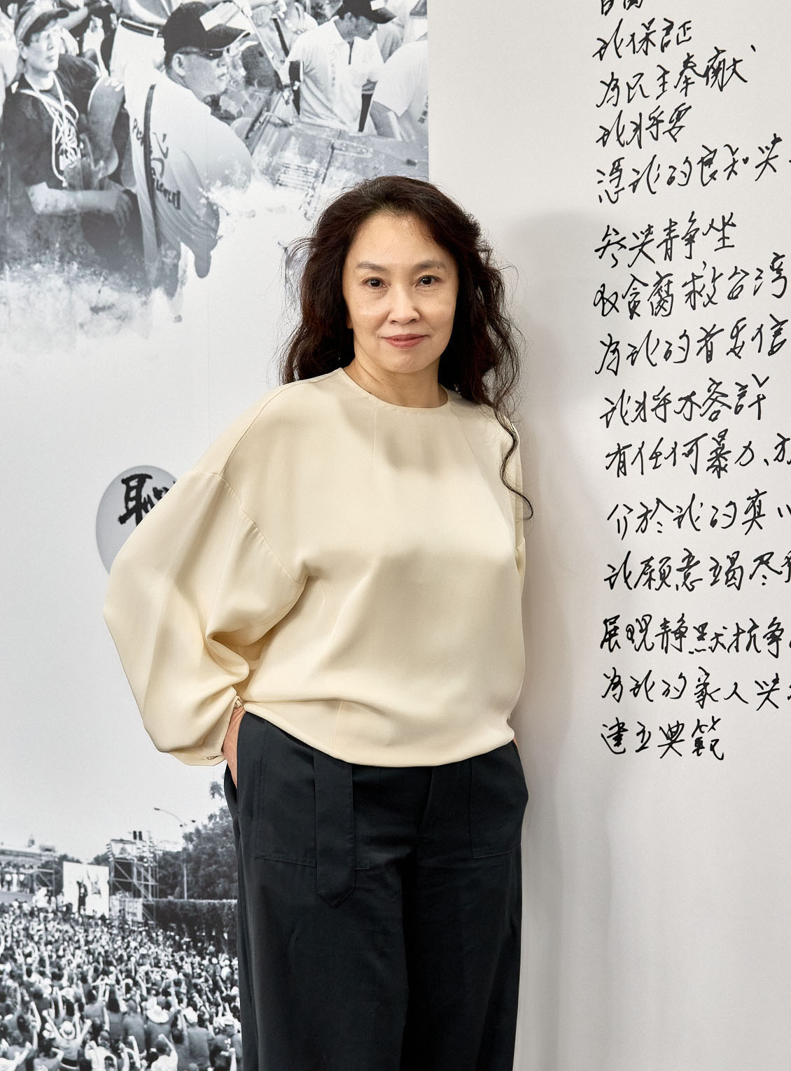 Woman in a beige blouse and dark pants stands beside a wall showing a large black-and-white crowd image and vertical Chinese script.