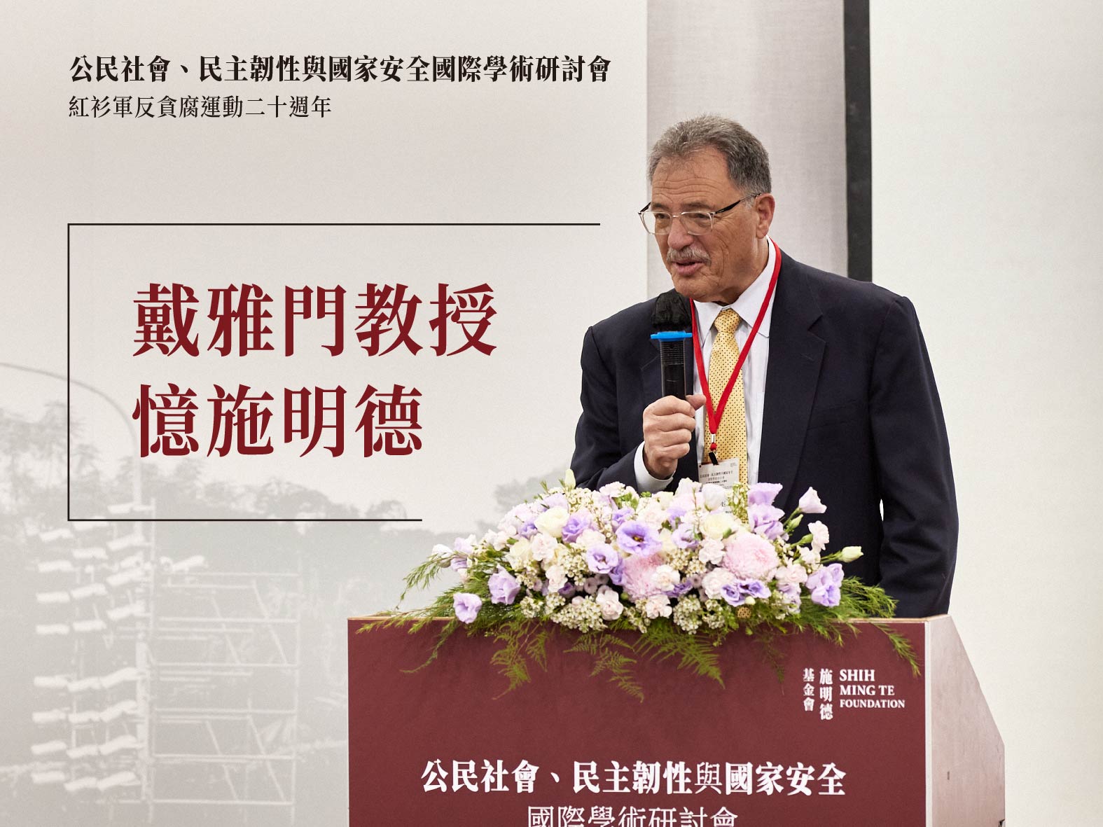 An older man in a suit speaks at a conference podium adorned with flowers, with Chinese text on a banner beside him and in the background laterally.